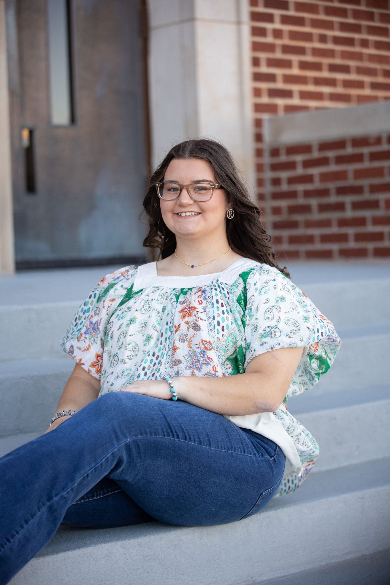 A person in a floral print top and blue jeans sits casually on concrete steps against a brick wall.