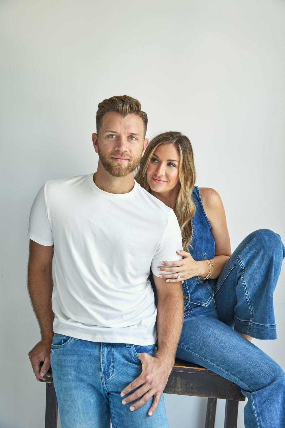 Casual couple in jeans and white shirts posing together against light background.