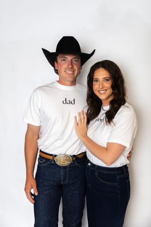 Couple wearing matching white t-shirts and blue jeans pose together against white background in western style.