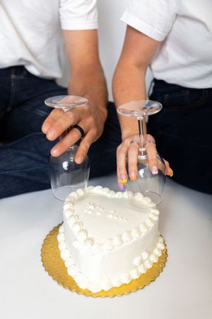 Hands holding wine glasses upside down over a heart-shaped white frosted cake on a gold board.