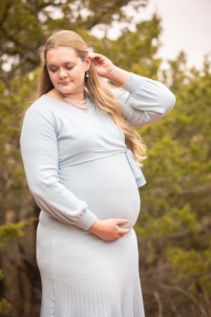 A person in a light blue sweater cradles their pregnant belly while posing outdoors against a natural backdrop.