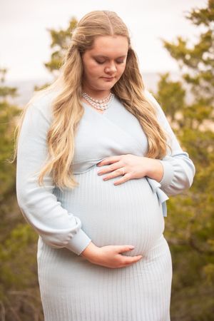 Pregnant woman in light blue sweater cradles her baby bump in outdoor maternity photoshoot at sunset.