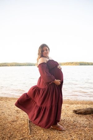 A person in a burgundy dress sits on a rocky beach shoreline at sunset, creating a serene coastal scene.