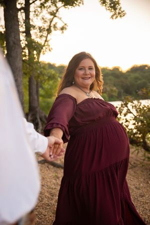 Person in burgundy dress poses outdoors at sunset with trees and water in background.