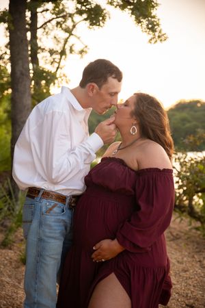 A romantic couple shares tender moments in a wooded area at sunset, with the person in burgundy wearing an off-shoulder dress.