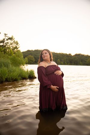 Two people in burgundy outfits embrace while standing in a lake at sunset with trees in the background.