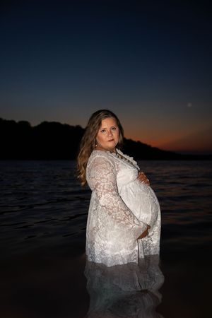 A pregnant woman in white lace dress poses in water at sunset with dark sky and trees in background.