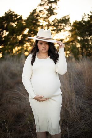 A person in a white sweater poses outdoors at sunset against a backdrop of dried grass and trees.