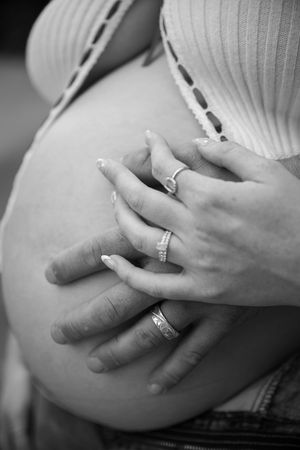 Black and white photo of hands with wedding rings resting on a pregnant belly in a white ribbed top.