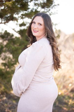 Woman in white sweater poses outdoors at sunset with mountains and evergreen trees in background.