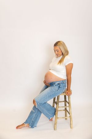 Person wearing light colored top and blue jeans poses on wooden stool against white background in artistic photo series.
