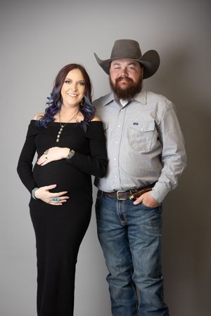 A couple poses together in a studio setting, with the man wearing a cowboy hat and the woman in a black dress.