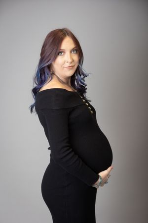 A woman in a black sweater dress poses during a maternity photo shoot against a gray backdrop.