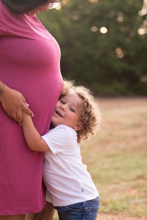 A child in a white t-shirt hugs a pregnant person wearing a pink shirt in an outdoor setting at sunset.
