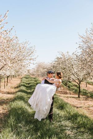 Playful couple dancing in white dress and casual wear among blooming orchard trees.