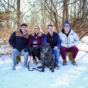 A group sits together with their black dog on a snowy winter day in the woods.