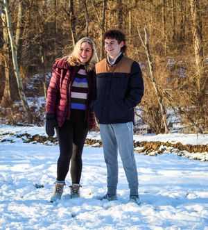 Two people walk together on a snowy winter path surrounded by bare trees and white ground.