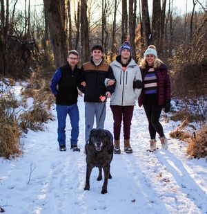 A group walks with their black dog on a snowy winter trail through bare trees in a forest setting.