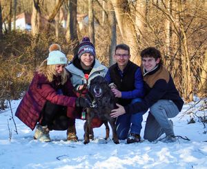 A winter group photo in the snow with a happy black dog surrounded by winter clothing.