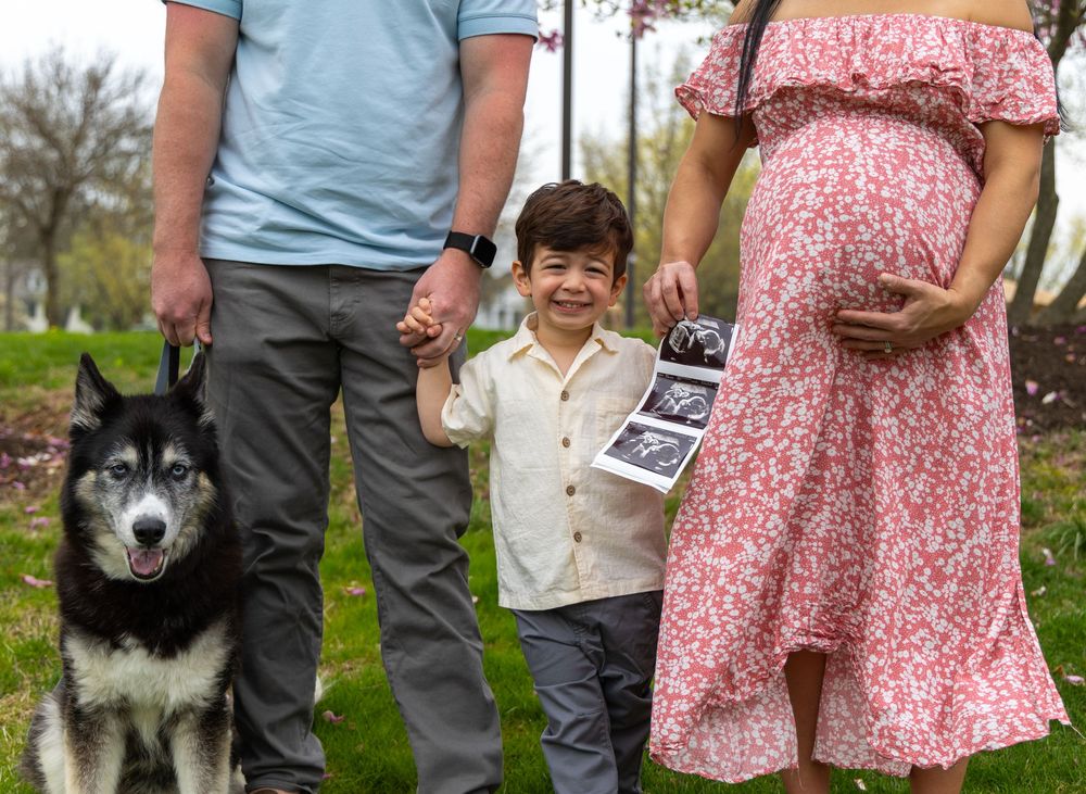 A family poses outdoors with their Alaskan Malamute dog wearing a pink floral dress and casual attire.
