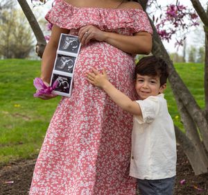 A pink floral maternity dress is showcased in a spring setting with blooming cherry trees in the background.