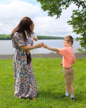 A woman in a floral dress interacts playfully with a child in a coral shirt by a lakeside.