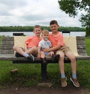 Three children in matching coral shirts sit together on a park bench near a lake on a cloudy day.