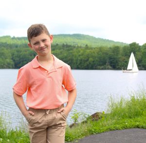 A person in coral shirt and khaki shorts poses near a serene lake with green hills in the background.