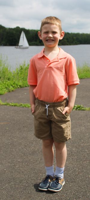 A child in a coral polo shirt and khaki shorts stands on a path by a lake with a sailboat in the background.