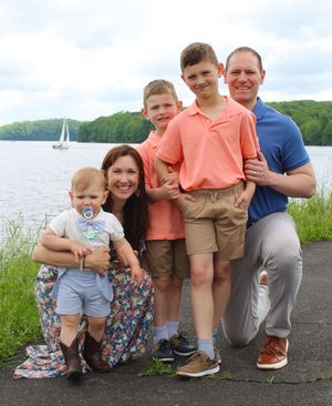 A family poses together on a stone wall near a lake with sailboats visible in the background.