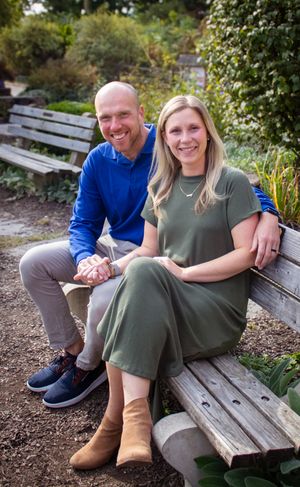 A couple sits together on a wooden bench in a garden setting wearing casual clothing.