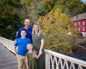 A group stands on a white wooden bridge with autumn foliage and a red building in the background.