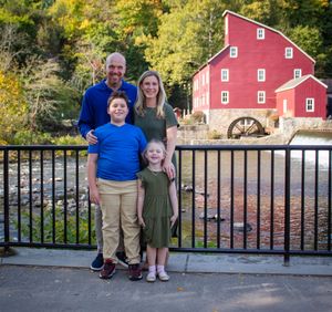 Family stands together on wooden bridge overlooking scenic pond with red barn in background on autumn day.