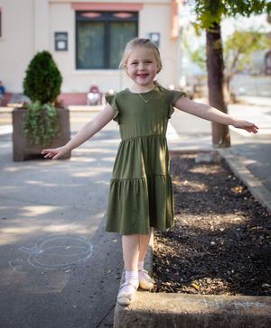 A child in an olive green dress twirls and plays on a sidewalk in front of a building on a sunny day.