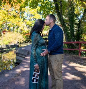A couple shares a tender moment on an autumn path while holding ultrasound photos in a forest setting.