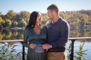 A couple in casual attire stands together by a serene lake surrounded by fall foliage.