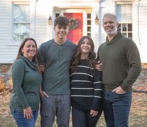 A group of four people wearing casual sweaters pose together for a family photo in front of a house with a red door.
