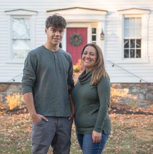Couple wearing matching dark green and gray outfits pose together in front of a house during autumn.