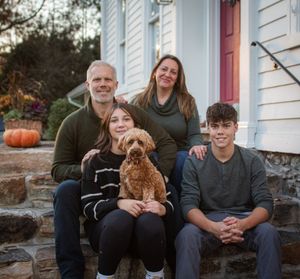 A family sits together on stone steps outside a white house with a small brown dog at sunset.