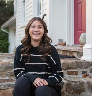 A person sits on stone steps outside a white building with a red door wearing a black and white striped sweater.
