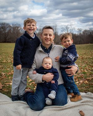 A family sits together on a gray blanket outdoors during autumn with fallen leaves scattered on the ground.