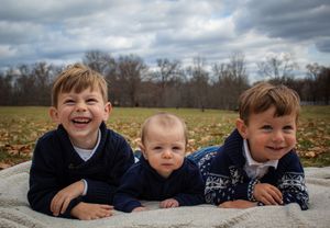 Family group lying on blanket outdoors with cloudy sky and autumn trees in background.