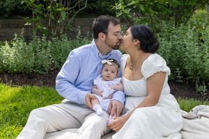 A couple kisses their baby girl while sitting on a blanket in a lush garden setting.