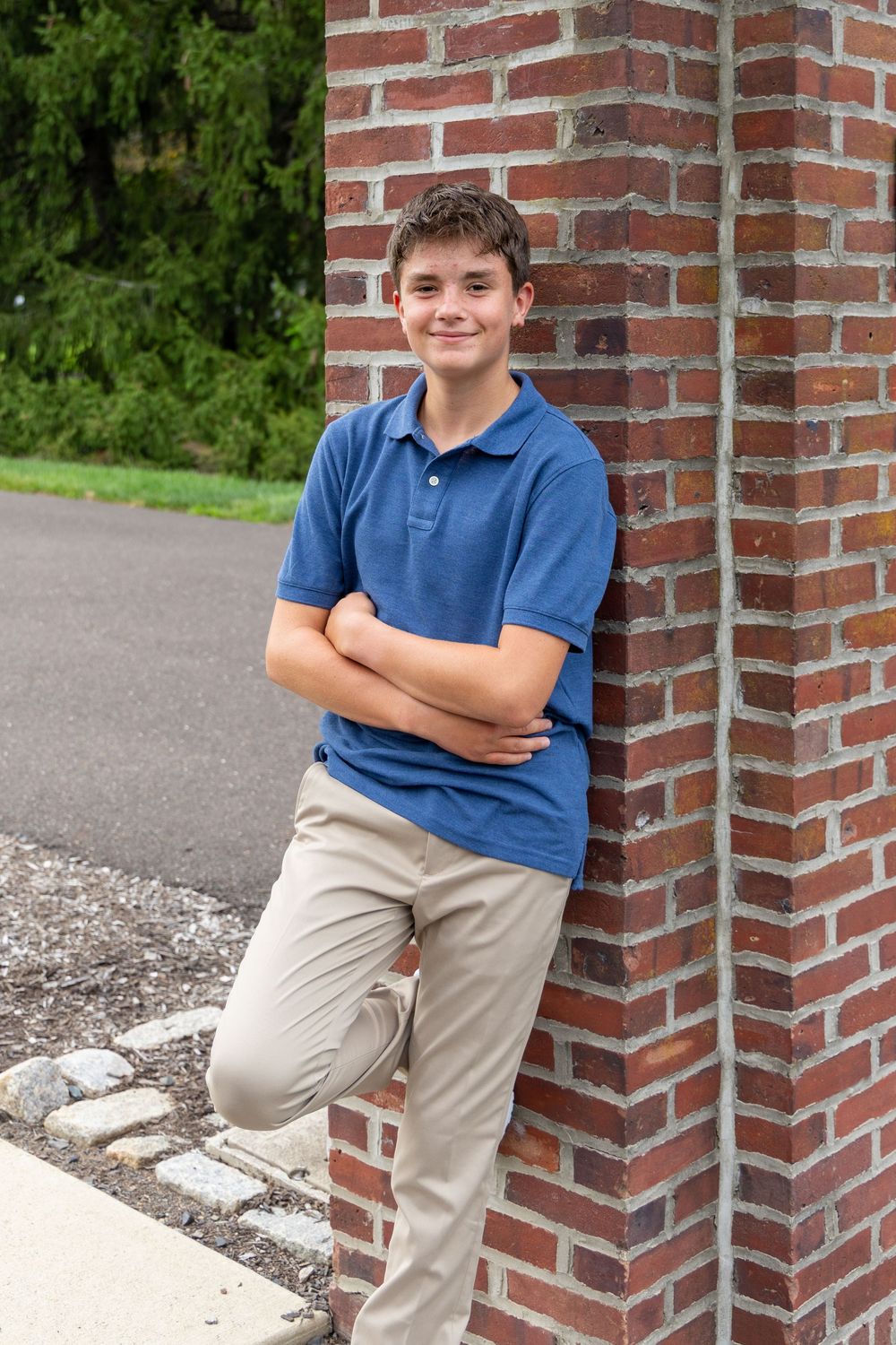 Teen boy in blue polo and khakis leans against brick pillar with arms crossed, smiling outdoors.