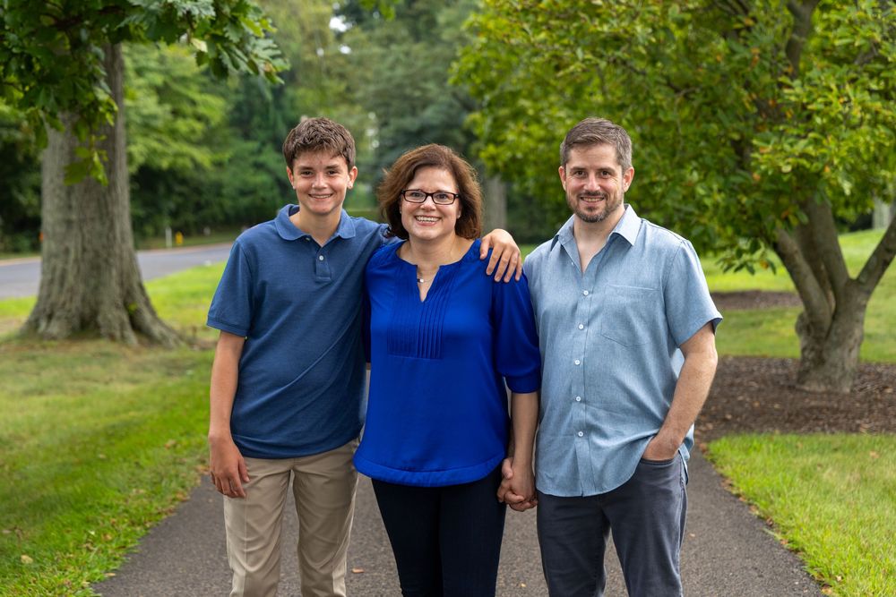 Family of three smiling together on a tree-lined path in a lush green park on a sunny day.