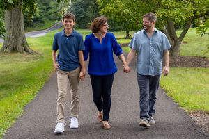 Family of three walking hand-in-hand on a tree-lined path, smiling outdoors on a sunny day.