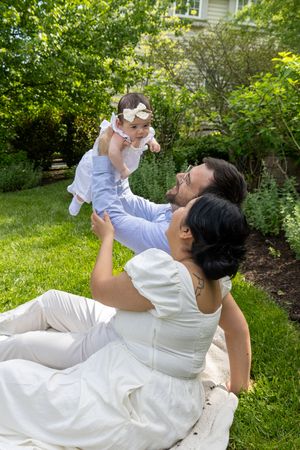 A happy couple sits on grass lifting their baby girl in the air in a lush green backyard.