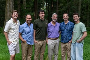 Six men smiling together outdoors in a wooded setting, wearing casual summer attire.
