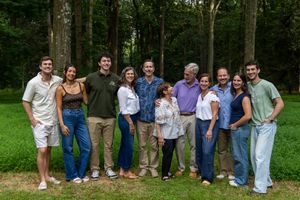 Large family group smiling together outdoors on green grass with tall trees in background.