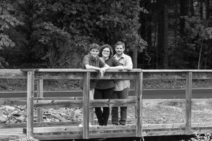 Black and white photo of a woman and two children smiling on a wooden bridge surrounded by trees.